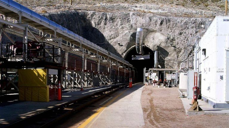 Široki ulaz u tunel uklesan u stjenovitu padinu, s metalnim konstrukcijama i opremom s obje strane. Ulaz je poduprt čeličnim gredama, a ventilacijski kanal se proteže u tunel. Staza koja vodi do ulaza ima tračnice. Vidljivi su razni strojevi.