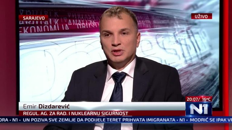 Muškarac u odijelu i kravati govori tijekom TV prijenosa uživo. Na ekranu se ispisuje tekst: "Emir Dizdarević, Regul. Ag. Za Rad. I Nuklearnu Sigurnost" i "N1". Pozadina prikazuje stiliziranu grafiku. Vijesti se pomiču na dnu s različitim naslovima.