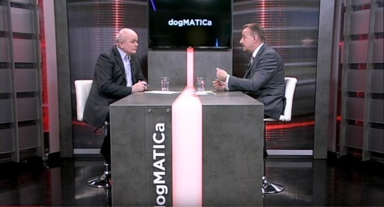 Dva muškarca u televizijskom studiju sjede jedan nasuprot drugoga za modernim pravokutnim stolom s tekstom "dogMATICa" sa strane. Čine se uključeni u raspravu, sa zaslonom iza njih koji prikazuje isti tekst. Studio ima crvene i crne akcente.