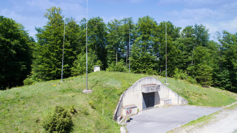 Travnati, zemljom prekriven vojni bunker s betonskim ulazom nalazi se među bujnim zelenim drvećem na sunčan dan. Antene su postavljene na stupovima oko bunkera, a na popločanom prostoru ispred se nalazi dio opreme i predmeta vidljivih u blizini ulaza.