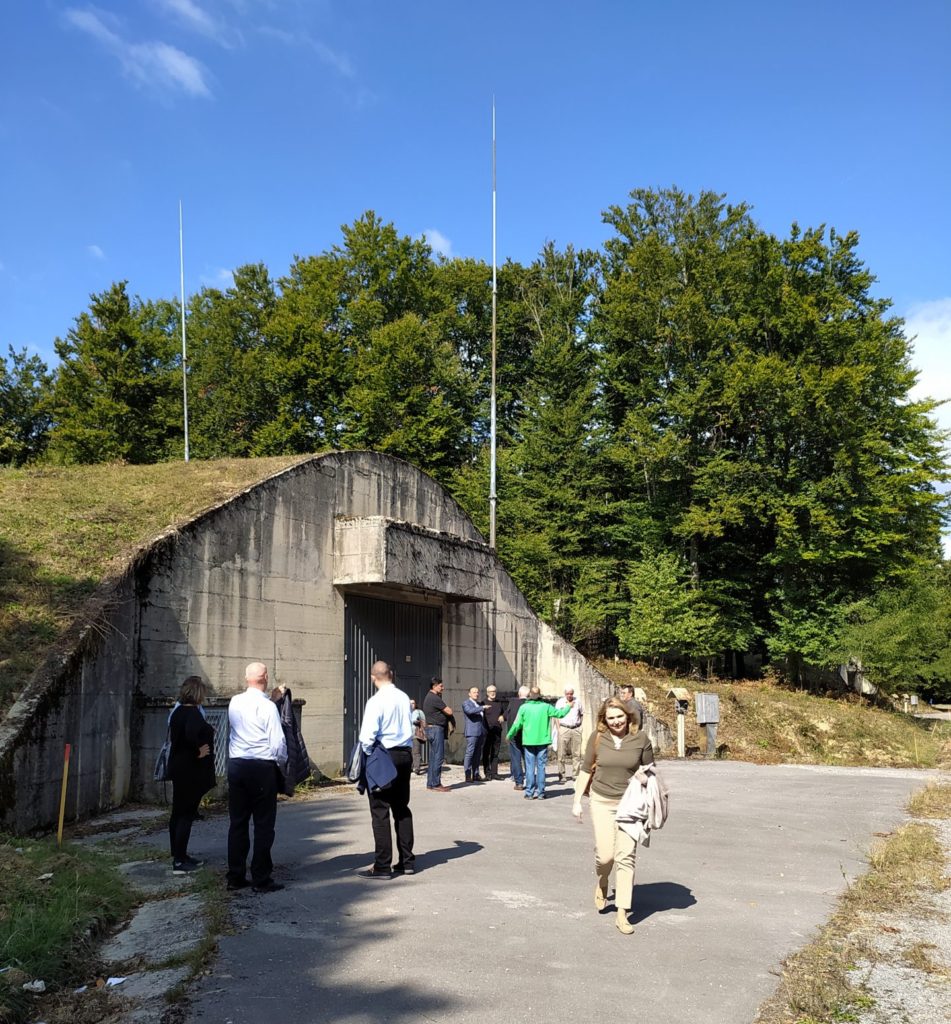 Grupa ljudi stoji i šeta blizu betonskog bunkera na sunčan dan. Bunker je djelomično prekriven travom i drvećem u pozadini. Neki pojedinci razgovaraju dok se drugi udaljavaju od ulaza u bunker.