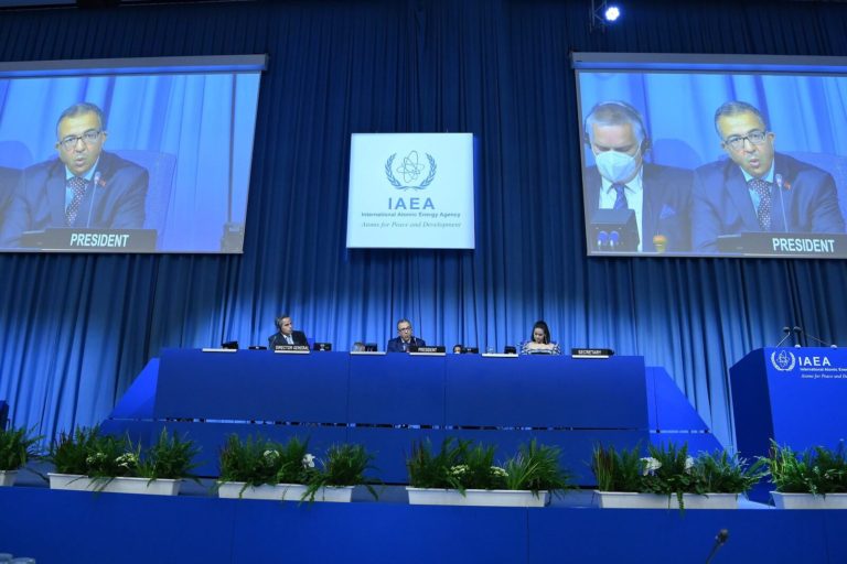 Konferencijska dvorana s velikom pozadinom s plavim zavjesama prikazuje amblem Međunarodne agencije za atomsku energiju (IAEA). Četvero ljudi sjedi za stolom na pozornici, s natpisom "Predsjednik" i dva velika ekrana iznad koji prikazuju predsjednika kako govori.