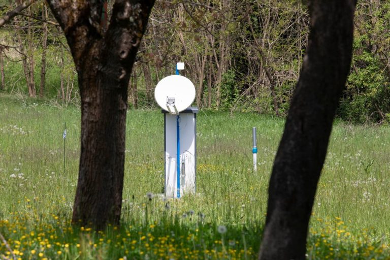 Satelitska antena i oprema meteorološke stanice stoje na bujnom travnatom polju okruženom drvećem. Antena i oprema postavljeni su na plavo-bijeli stup, s vidljivim raznim senzorima i žicama. Jarko žuto i bijelo poljsko cvijeće prošarano je livadom.