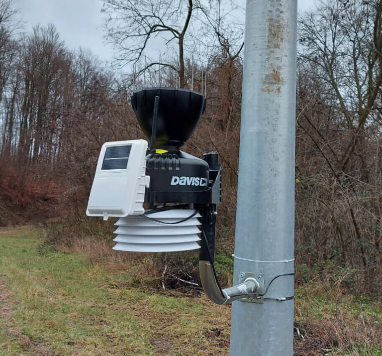 Meteorološka stanica postavljena na metalni stup na otvorenom, opremljena kišomjerom, solarnom pločom i raznim senzorima. Stanica je smještena na travnatom području s drvećem i golim granama u pozadini. Oblačno nebo ukazuje na oblačno vrijeme.