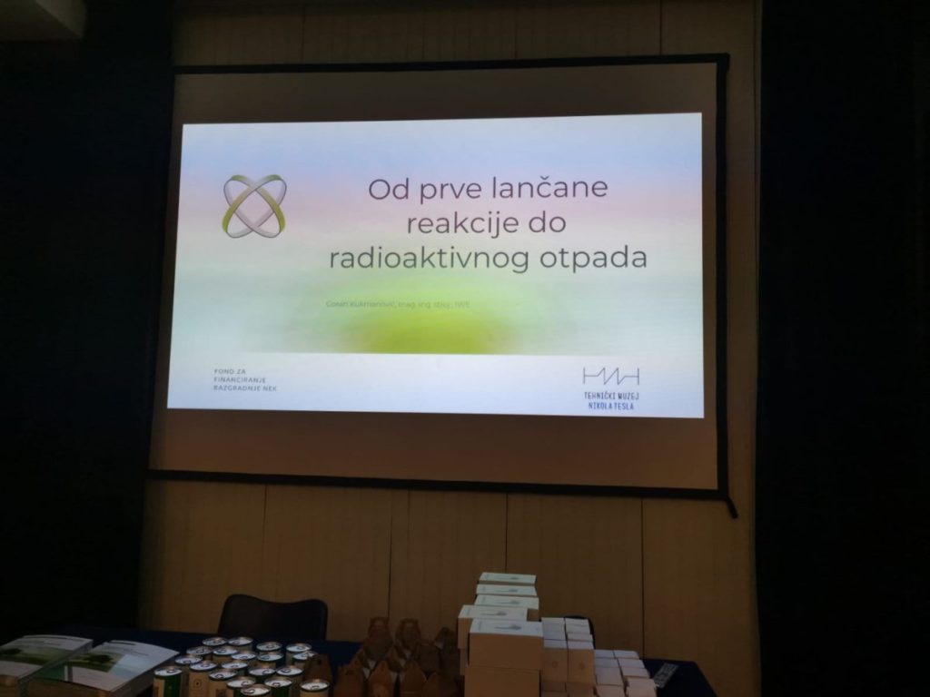 Prezentacijski zaslon s naslovom na hrvatskom jeziku: "Od prve lančane reakcije do radioaktivnog otpada" što u prijevodu znači "Od prve lančane reakcije do radioaktivnog otpada". Razne kutije i predmeti postavljeni su na stol ispred ekrana.