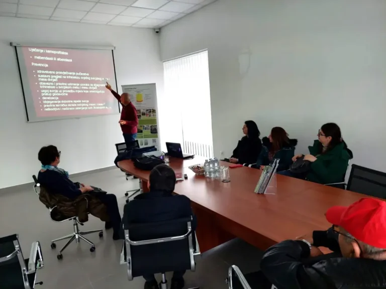 Osoba koja stoji pored platna projektora održava prezentaciju grupi ljudi koji sjede oko velikog stola u svijetloj konferencijskoj sobi. Slajd na ekranu sadrži tekst i popis s grafičkim oznakama na poljskom. Na stolu je nekoliko dokumenata i elektronika.