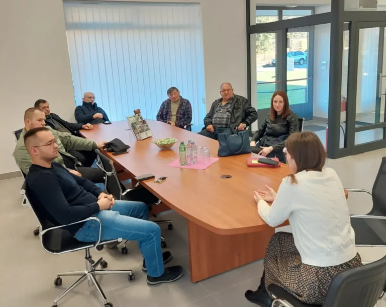 Osam ljudi sjedi oko konferencijskog stola u uredskom okruženju. Žena sprijeda razgovara sa skupinom. Razni predmeti, uključujući boce s vodom i mape, stavljaju se na stol. Veliki prozor u pozadini prikazuje automobile parkirane vani.