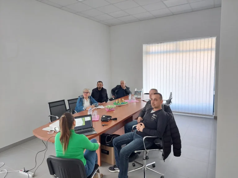 Grupa od šest ljudi, pet muškaraca i jedna žena, sjedi oko pravokutnog konferencijskog stola u uredu. Jedna žena sjedi za prijenosnim računalom leđima okrenuta kameri. Soba ima bijele zidove i veliki prozor s okomitim roletama. Na stolu su razni predmeti.