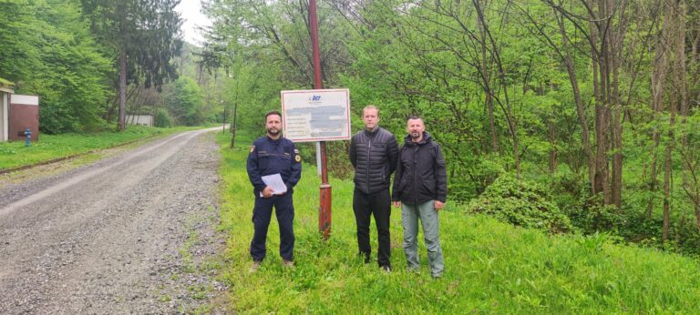 Tri muškarca stoje pored znaka u bujnom, zelenom šumskom području. Uz njih prolazi makadamska cesta. Znak sadrži tekst i logotipe i montiran je na visoki stup. Muškarci su ležerno odjeveni i gledaju prema kameri.