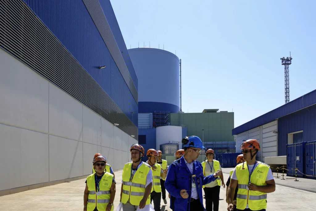 Grupa ljudi u žutim sigurnosnim prslucima i kacigama hoda ispred industrijskog pogona s velikim plavim i sivim zgradama i spremnicima. Čini se da jedna osoba u plavoj uniformi vodi skupinu, a svi izgledaju uključeni u razgovor.