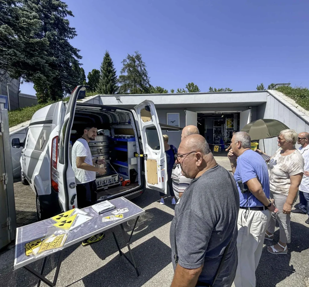 Grupa ljudi okuplja se oko otvorenog stražnjeg dijela bijelog kombija, gdje muškarac pokazuje ili objašnjava opremu pohranjenu unutra. Čini se da je mjesto vanjsko područje s drvećem i zgradom u pozadini. U prvom planu je stol s pamfletima.