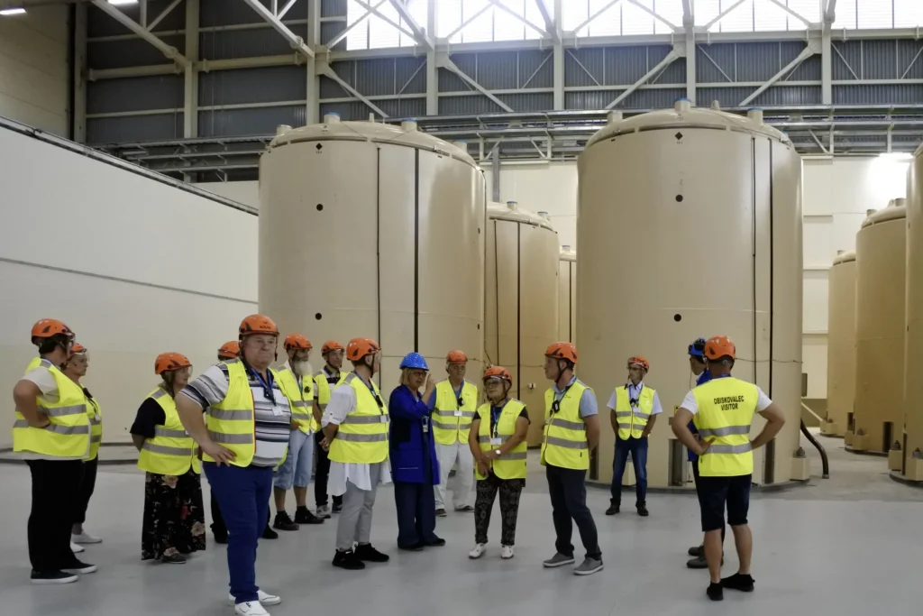 Grupa ljudi sa zaštitnim prslucima i kacigama okupljena je unutar industrijskog pogona, stojeći ispred velikih, cilindričnih spremnika. Čini se da slušaju osobu u plavoj košulji koja govori ili vodi turneju.