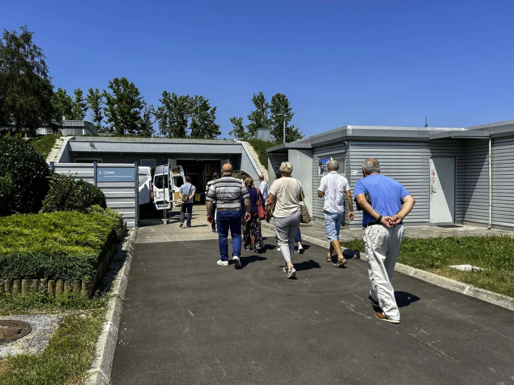 Grupa ljudi hoda prema zgradi sa sivim rolo vratima pod vedrim plavim nebom. Bijeli kombi je parkiran kraj jednih otvorenih vrata. Staza je s jedne strane okružena zelenilom i grmljem. Čini se da su ljudi ležerno odjeveni.