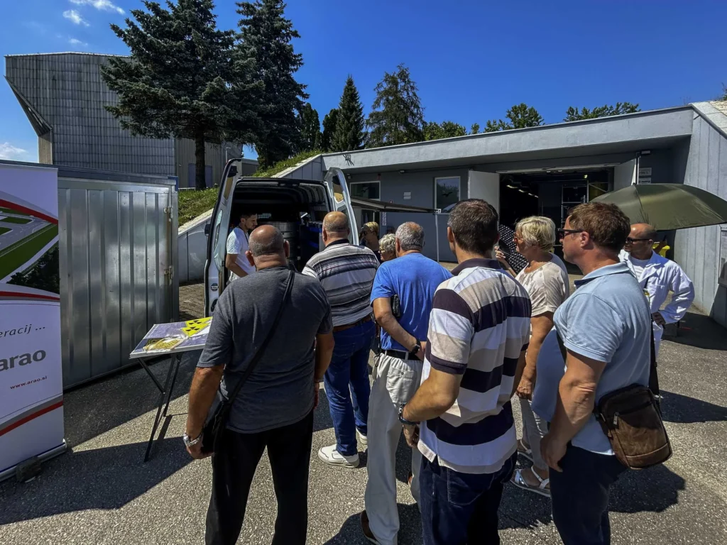 Grupa ljudi stoji u redu vani na sunčanom danu, čekajući iza kombija. Neki drže pamflete, au pozadini su stabla i moderna zgrada. Na lijevoj strani slike djelomično se vidi znak ili transparent.